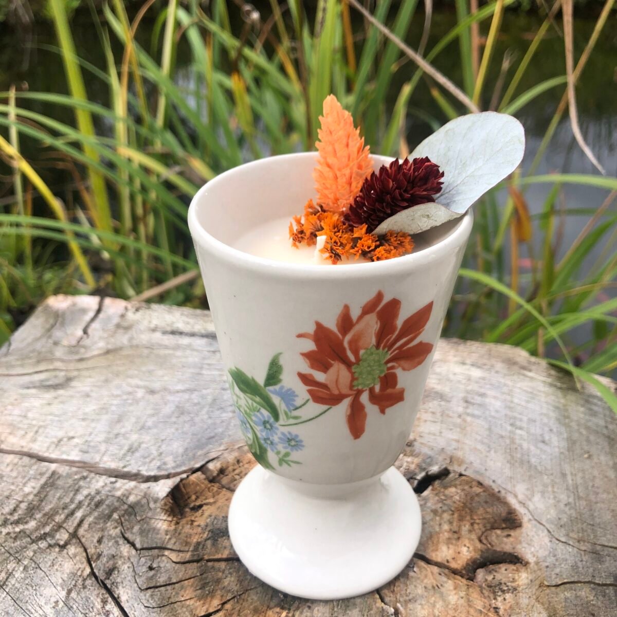Bougie artisanale dans un mazagrans vintage crème avec une fleur dans les tons orangers avec des fleurs séchées sur un fond naturel et aquatique
